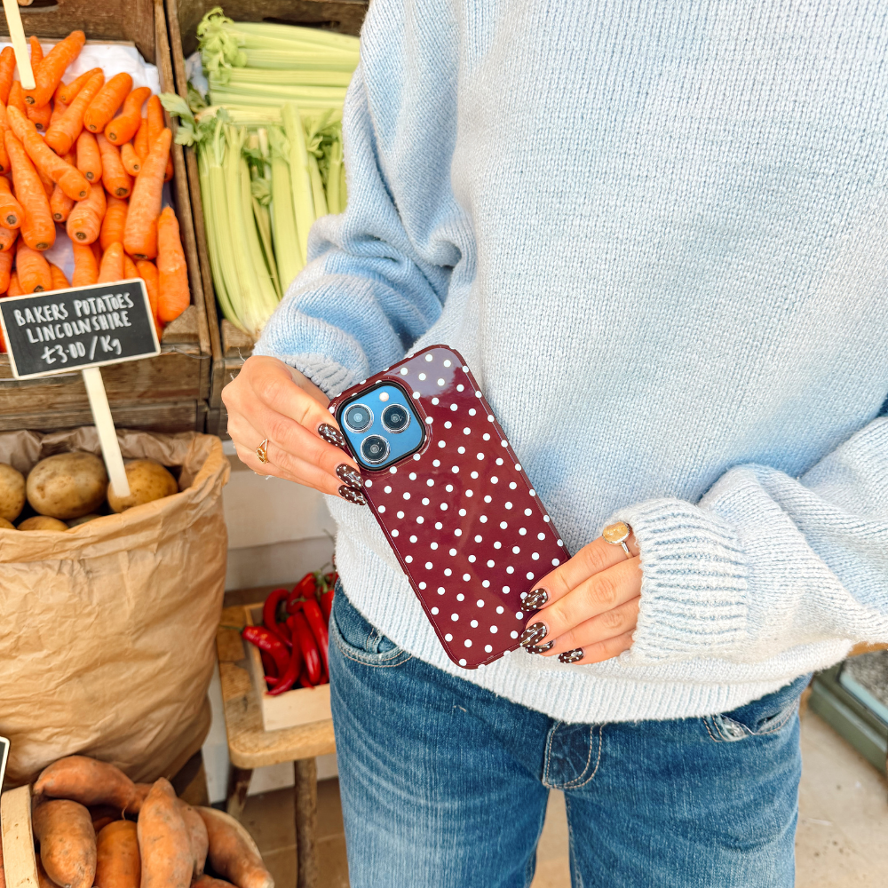 Girl holding her brown and blue polka dot tough phone case wearing a blue sweater with matching nails next to vegetables stacked up