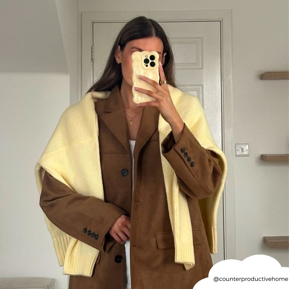 Woman holds a butter yellow 3D bubble phone case, taking a mirror selfie in a brown blazer.