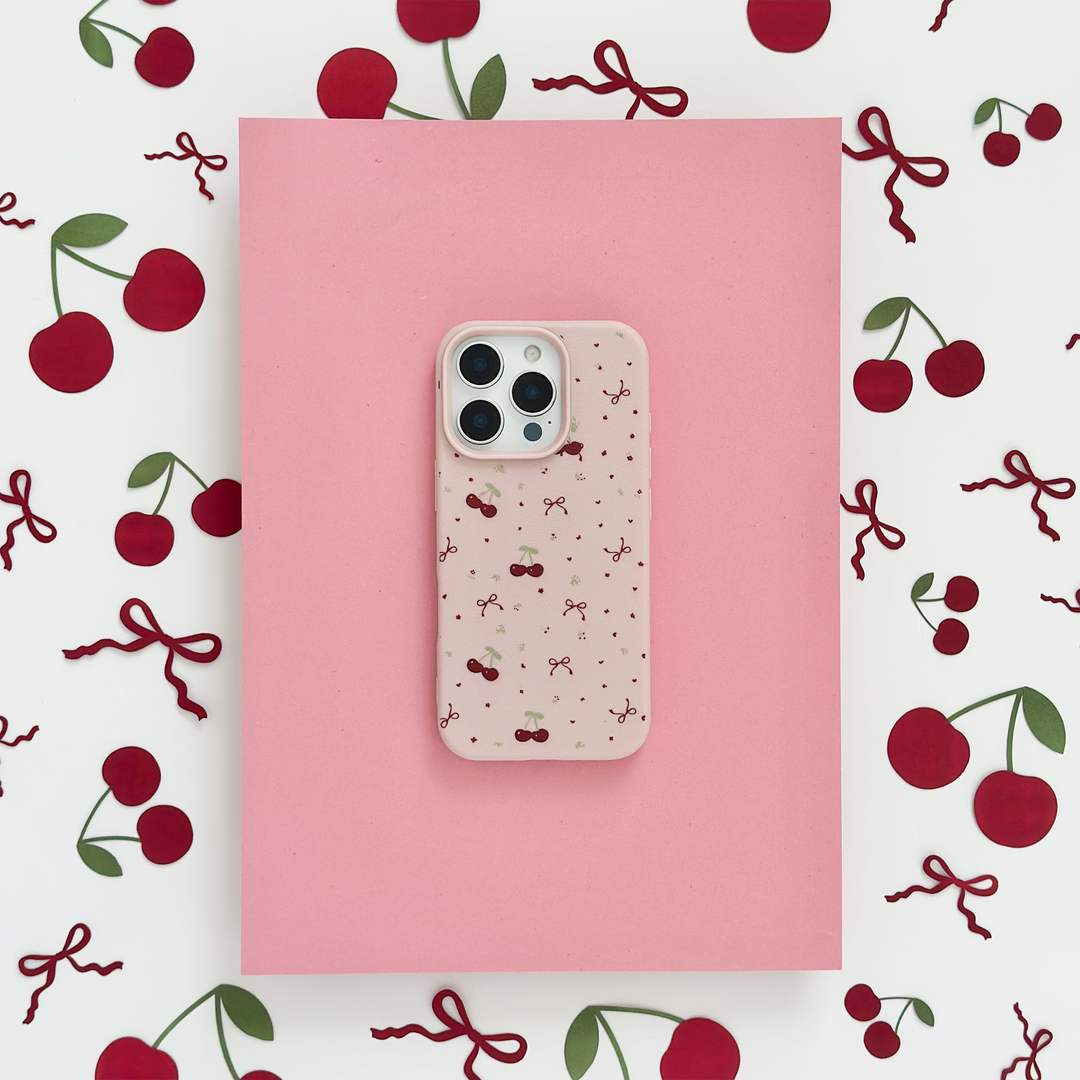 A smartphone with a pink, cherry-patterned case is placed on a pink paper sheet. The background is decorated with red cherries and bows scattered around.
