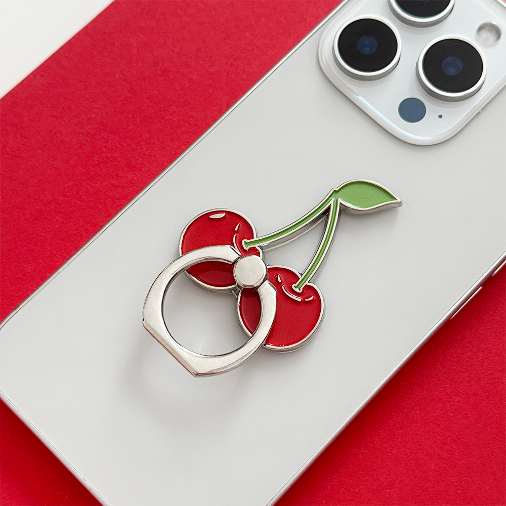 A silver smartphone with a cherry-shaped metal phone ring attached to the back, resting on a red and white background. The phone ring features two red cherries with green stems and leaf.