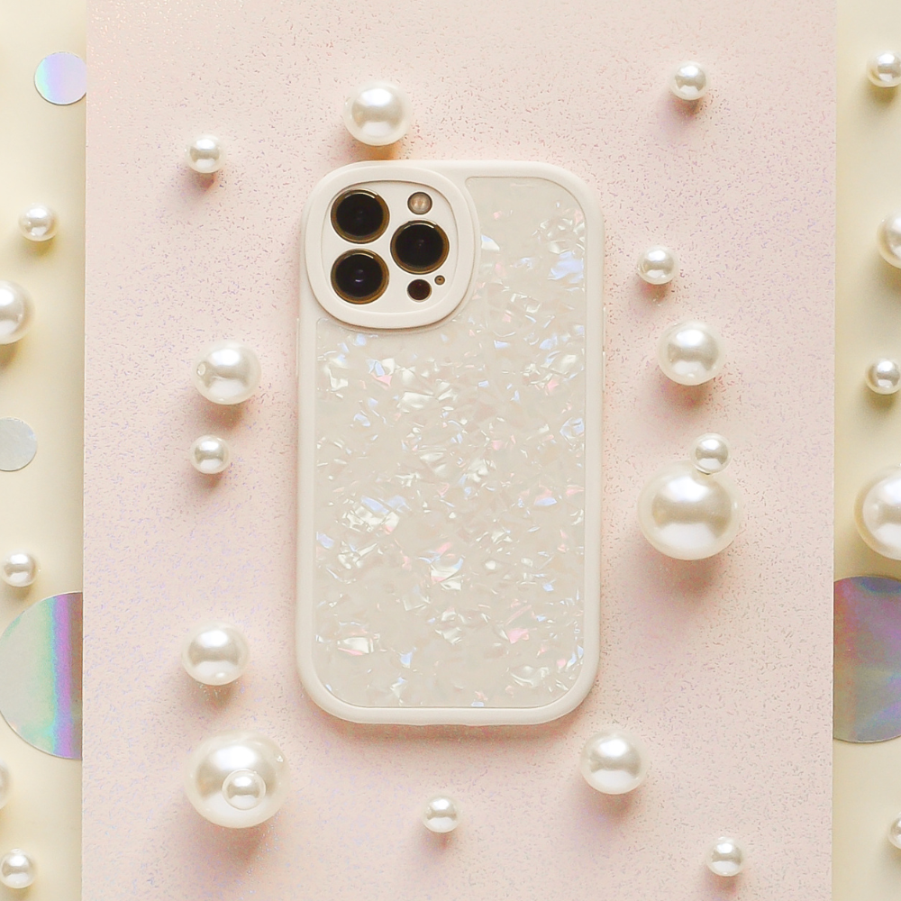 A white smartphone with a pearlescent, shimmery case is placed on a pastel pink, textured surface, surrounded by various sizes of white and iridescent pearls.
