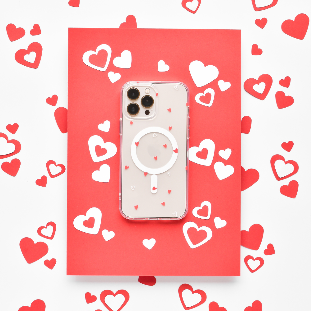 A smartphone with a high-quality Coconut Lane Clear MagSafe® Phone Case - Cute Hearts, adorned with tiny red hearts, is placed on a red card. The setup is surrounded by scattered red and white heart-shaped cutouts, creating a romantic and festive theme.