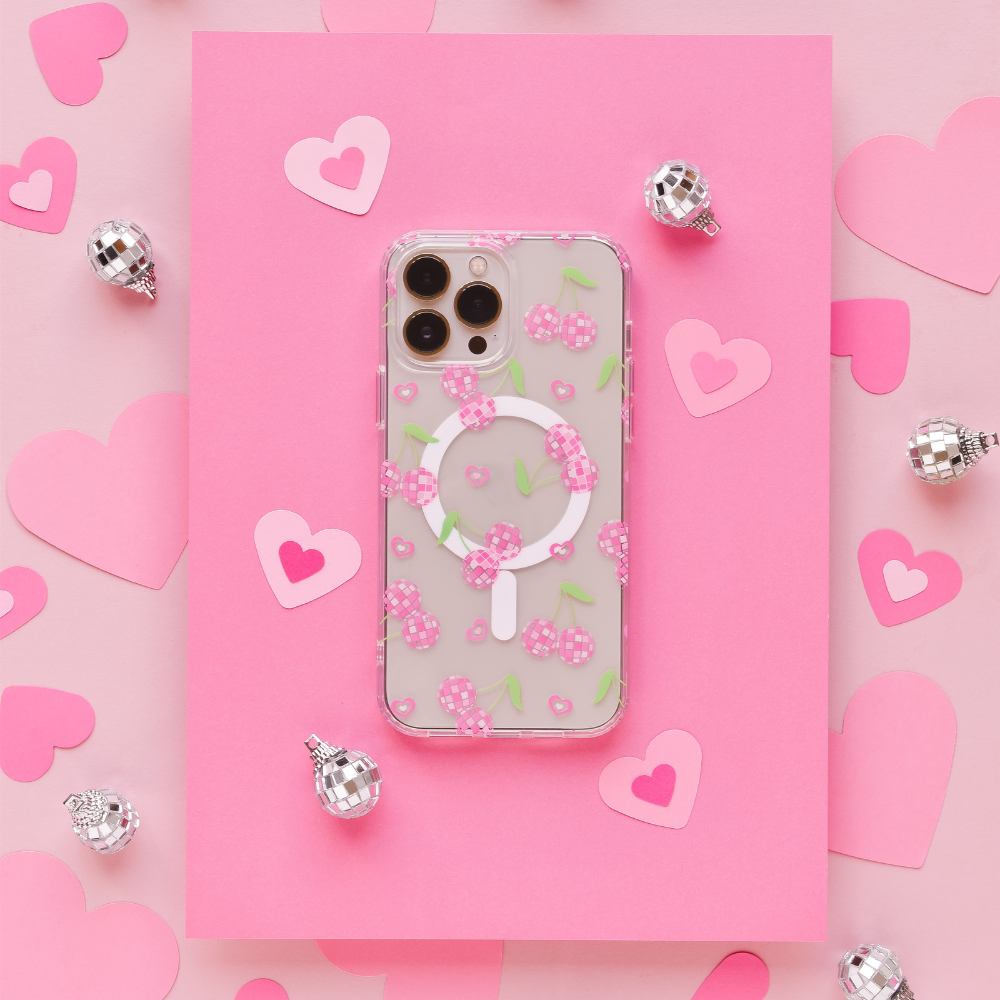A smartphone with a Coconut Lane Clear MagSafe® Phone Case in Disco Cherries design is placed on pink paper with heart cutouts. Silver disco ball ornaments are scattered around, enhancing the playful and romantic theme.