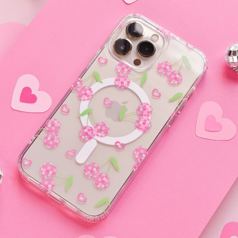 A smartphone equipped with the Coconut Lane Clear MagSafe® Phone Case - Disco Cherries, featuring pink checkered cherries, green leaves, and a white circular grip, is placed on a pink surface. Surrounding the phone are small pink heart-shaped cutouts and two tiny silver disco balls.