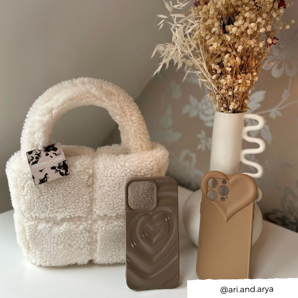 A cozy setup features a fluffy white handbag, two stylish phone cases—one Coconut Lane Melting Heart Phone Case in chocolate brown with a 3D melting heart effect, the other beige with a heart shape for the camera lens—and a white vase holding dried flowers. The image source, "@ari.and.arya," is noted in the bottom right corner.