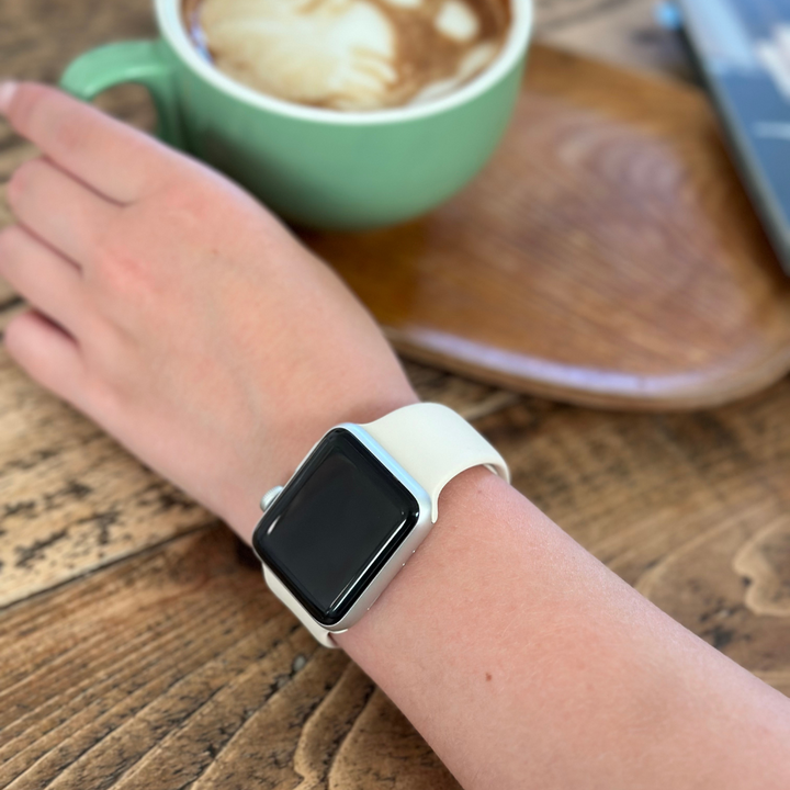A person's hand with an Apple Watch featuring the NAKD Apple Watch Strap in Almond White from Coconut Lane rests on a wooden table. Next to the hand is a green cup filled with latte, showcasing latte art on the foam. A wooden saucer and part of a book or magazine are also visible on the table.