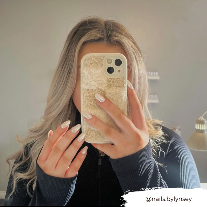 A person with long, wavy blonde hair takes a mirror selfie, covering their face with a phone adorned in Coconut Lane's Cosy Teddy Phone Case in Vanilla Biscuit. They are wearing a black top, and their nails are painted white. The handle "@nails.bylynsey" is displayed in the bottom right corner.