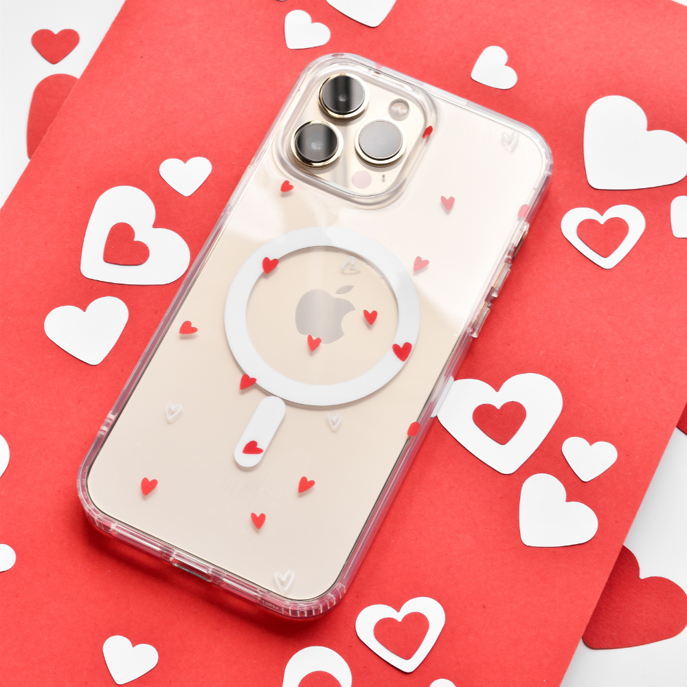 Placed on a red and white background with scattered heart-shaped cutouts, the Clear MagSafe® Phone Case - Cute Hearts by Coconut Lane offers high-quality protection while showcasing the phone's camera lenses, MagSafe® technology, and Apple logo through its transparent cover adorned with small red hearts.