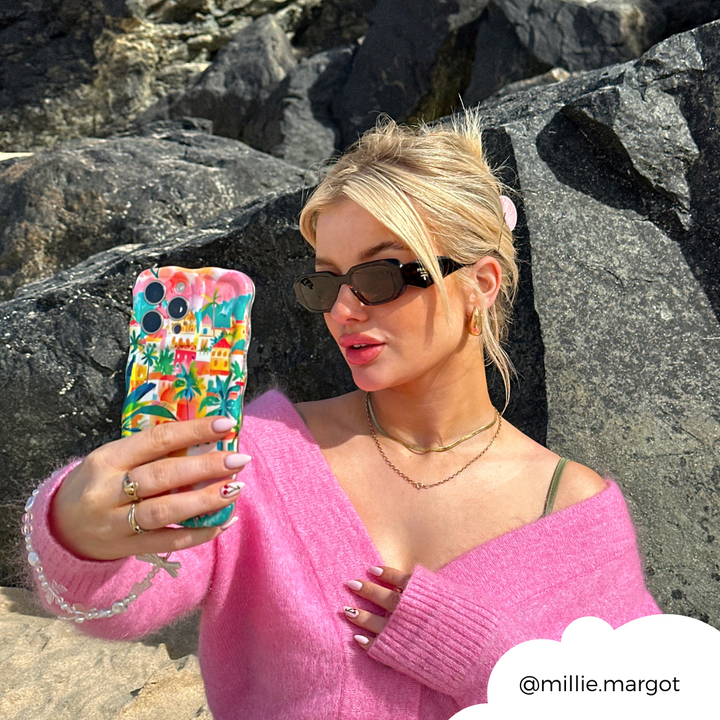 A blonde woman wearing sunglasses and a pink sweater is taking a selfie with her smartphone, which features the extra protective "Wavy Phone Case - Portofino Dreams" by Coconut Lane. She stands in front of large rocks, and her hair is tied back. Her Instagram handle "@millie.margot" is displayed in the lower right corner.