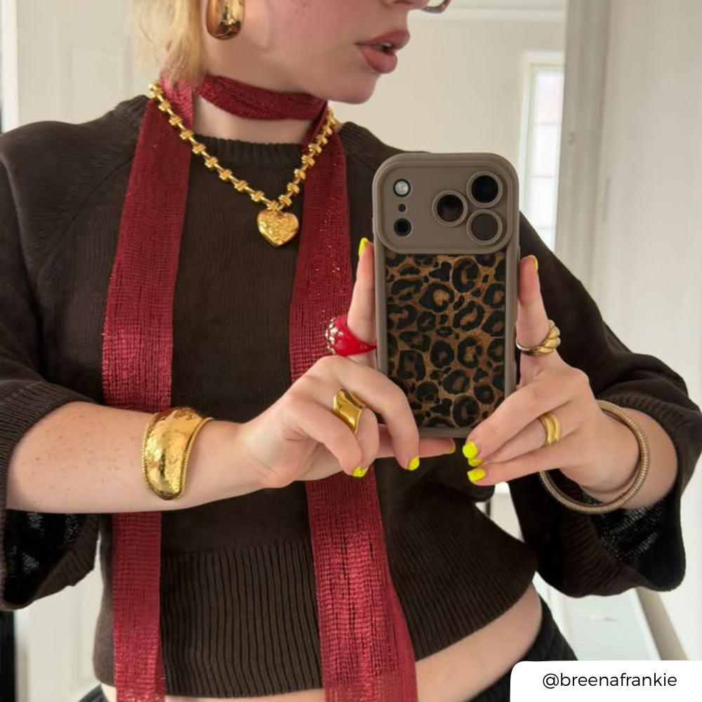 Woman taking a mirror selfie. She is wearing a chocolate brown jumper, accessorised with gold jewellery, a red mesh scarf. She is holding an iphone in a mocha leopard print phone case Her nails are painted yellow. 