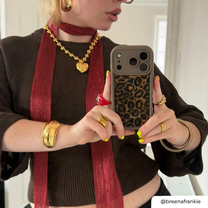 Woman taking a mirror selfie. She is wearing a chocolate brown jumper, accessorised with gold jewellery, a red mesh scarf. She is holding an iphone in a mocha leopard print phone case Her nails are painted yellow. 