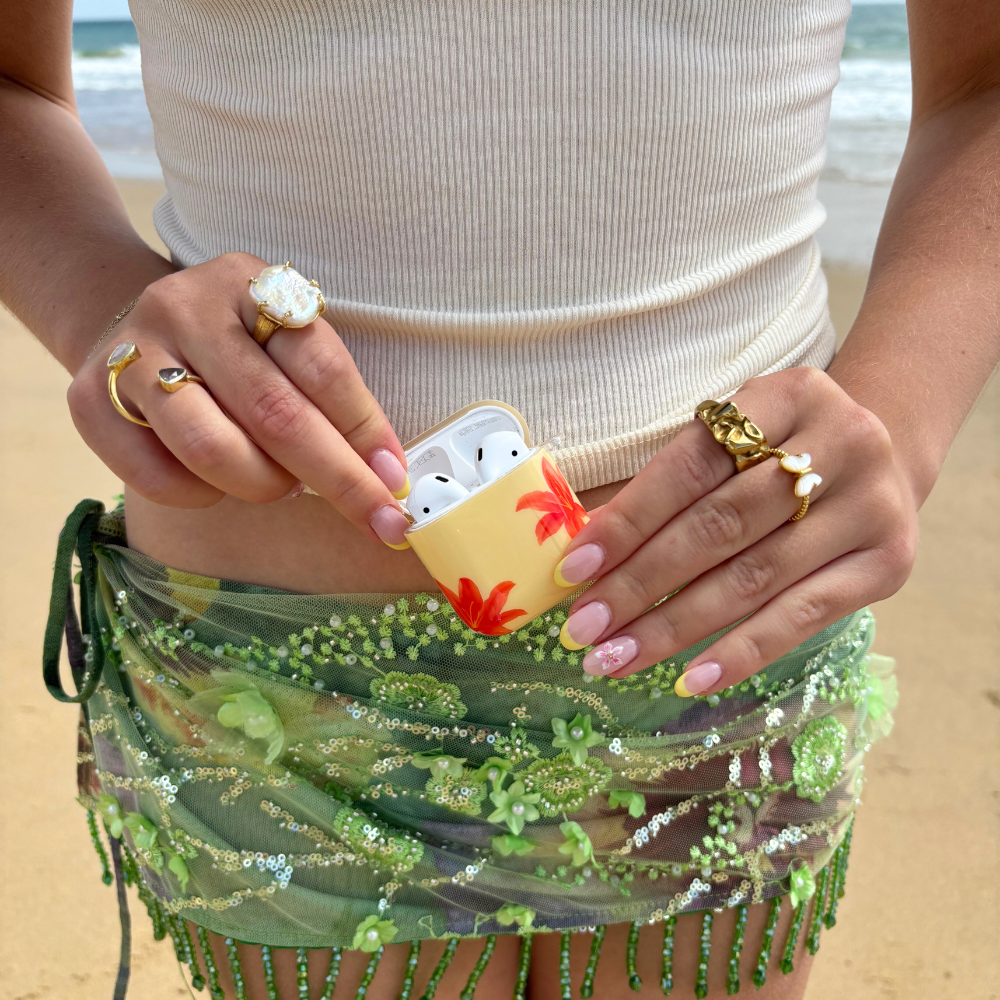 Person in white tank and green sarong holding a floral Coconut Lane AirPods case on a sandy beach.