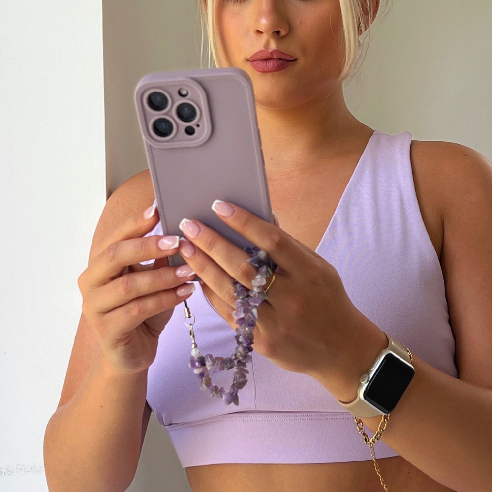A person with blonde hair wearing a light purple sports bra from the NAKD spring collection takes a photo with a lavender-colored iPhone. They have light pink nails, and their phone is protected by the Coconut Lane NAKD Phone Case in Lilac, which features an amethyst beaded wrist strap for Full Camera Protection. They are also wearing a smartwatch on their left wrist.