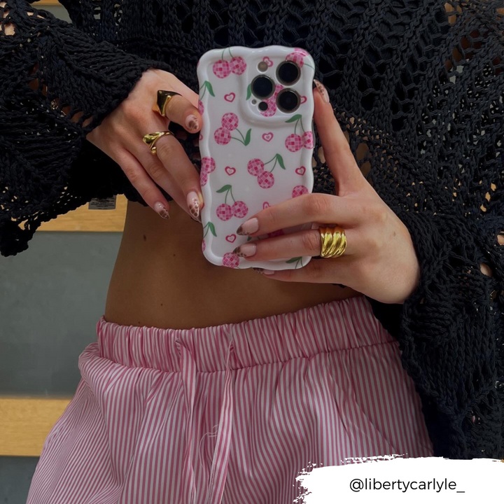 A person wearing a black crochet top and pink striped pants holds up a phone protected by the Wavy Phone Case - Disco Cherries from Coconut Lane. Their nails are adorned with nail art, and they are accessorized with several gold rings. The Instagram handle "@libertycarlyle_" is written at the bottom right.