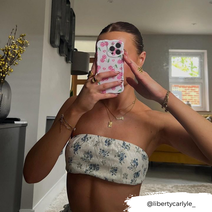 A person with slicked-back hair takes a mirror selfie, their face hidden by a phone adorned with Coconut Lane's Wavy Phone Case - Disco Cherries. They are dressed in a strapless, floral-patterned top. The room features modern decor and a large mirror. The Instagram handle @libertycarlyle_ is visible.