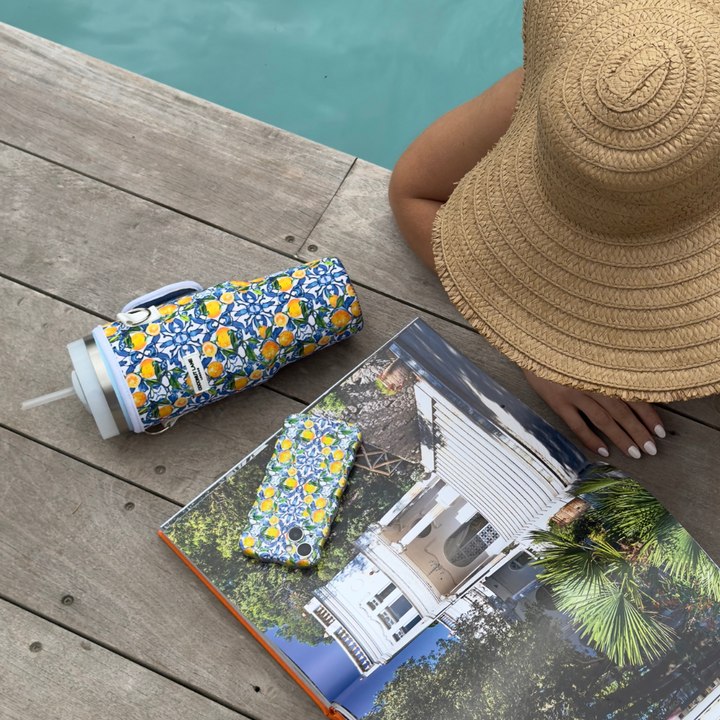 A person wearing a wide-brimmed straw hat and white nail polish sits poolside, with a floral-patterned Stanley Quencher travel mug, a Coconut Lane Tumbler Carry Case - Amalfi Lemon, a matching phone case, and an open book displaying images of a white house with greenery on a wooden deck surface.