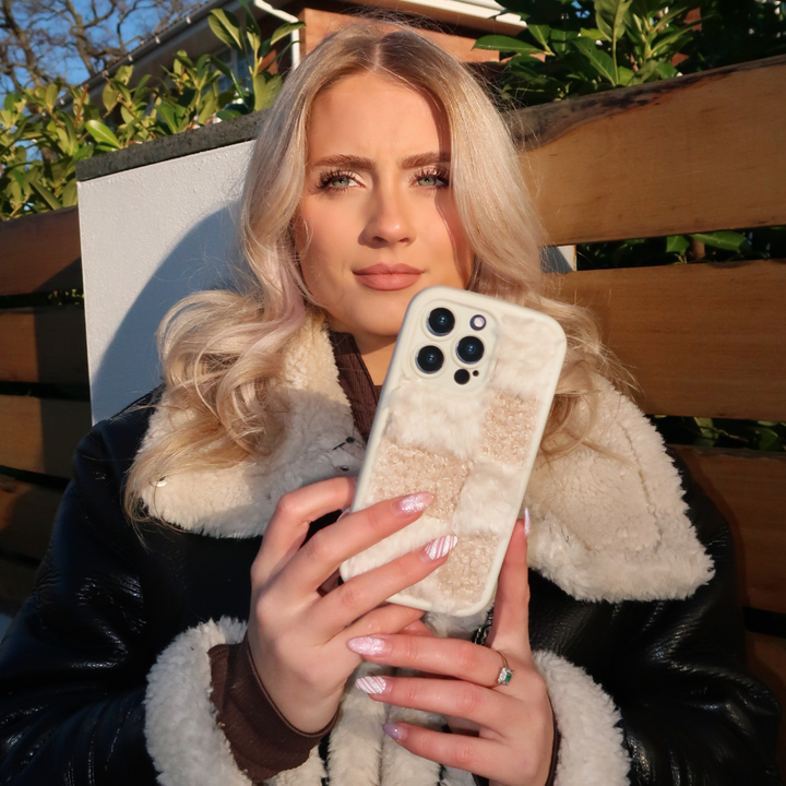 A young woman stands outdoors holding up a Coconut Lane Cosy Teddy Phone Case in Vanilla Biscuit. She has long, wavy blonde hair and is wearing a black jacket with a fluffy, light-colored inner lining. The background shows greenery and a brown wooden fence.