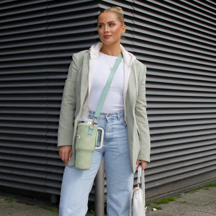A woman stands in front of a corrugated metal wall, dressed in a light green blazer, a white top, and light blue jeans. Carrying her green shoulder bag along with a white handbag, she confidently holds Coconut Lane's Tumbler Carry Case - Sage Heart in her free hand, with her hair styled back.