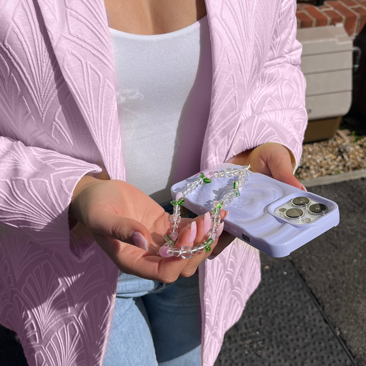 A person wearing a light pink textured blazer over a white top is holding a smartphone with a lavender case in one hand and the Coconut Lane Beaded Phone Strap - Cute Tulips in the other. The background includes a partial view of a brick house and some outdoor flooring.