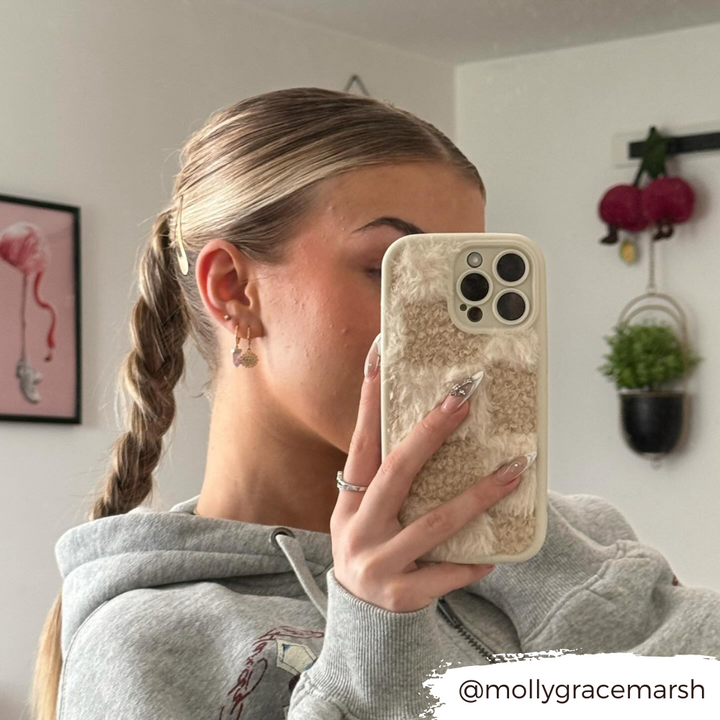 A person with a braided hairstyle takes a mirror selfie, showcasing the back of their phone adorned with the Cosy Teddy Phone Case - Vanilla Biscuit by Coconut Lane in beige and white checks. They are wearing a gray hoodie in a room decorated with various items, including a painting and hanging decor. The text "@mollygracemarsh" is on the image.