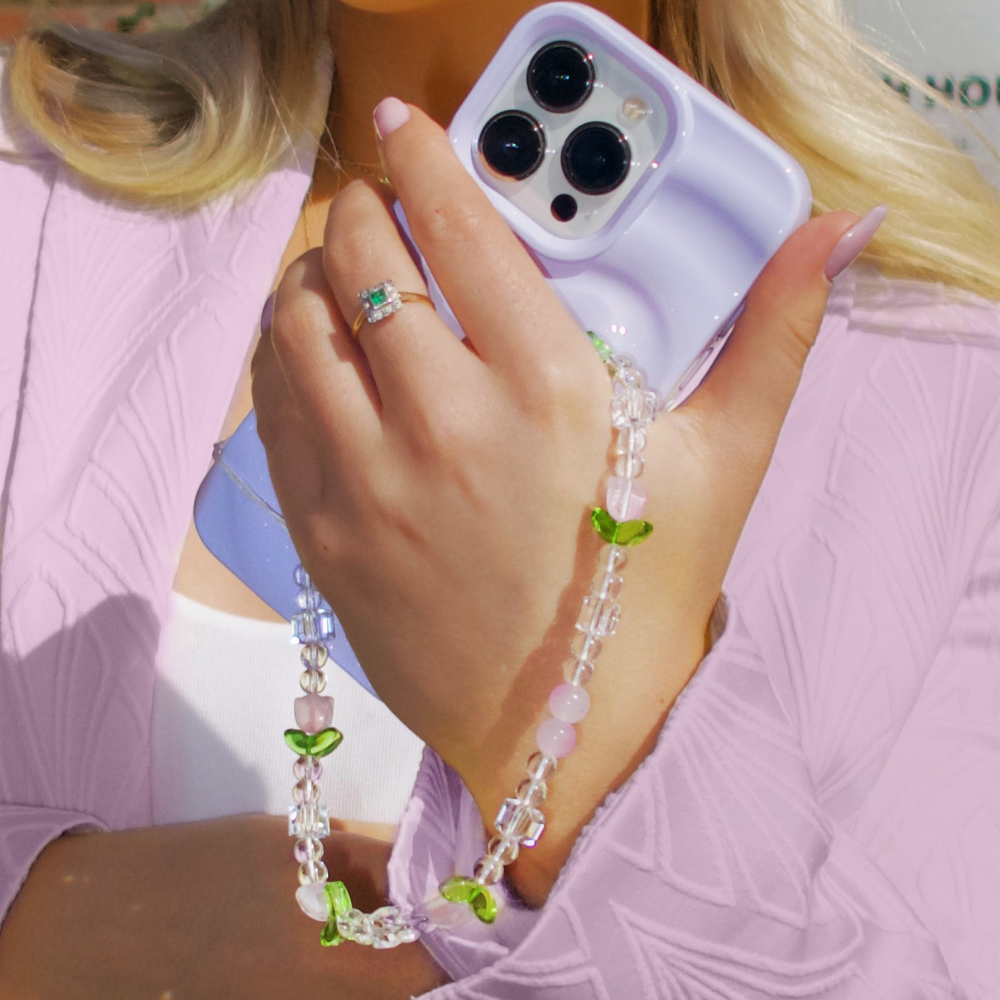 A person with long blonde hair holds a smartphone with a lavender case, featuring the Coconut Lane Beaded Phone Strap - Cute Tulips, adorned with green leaf accents. They wear a delicate ring with a green gemstone on their ring finger and a pastel purple jacket over a white top, exuding an effortlessly chic vibe.