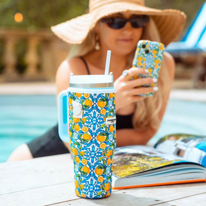 A person wearing a wide-brimmed straw hat, sunglasses, and a black swimsuit is sitting by a pool. They are using a smartphone with a decorative case, and there is a Coconut Lane Tumbler Carry Case - Amalfi Lemon next to an open magazine in the foreground.