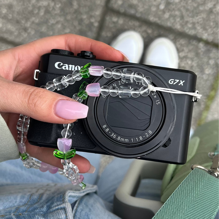 A hand with manicured nails holds a black Canon G7X camera, adorned with Coconut Lane's Beaded Phone Strap - Cute Tulips featuring pink, clear, and green beads. The background shows a stone pavement and a glimpse of the person's white shoes and part of a green bag.