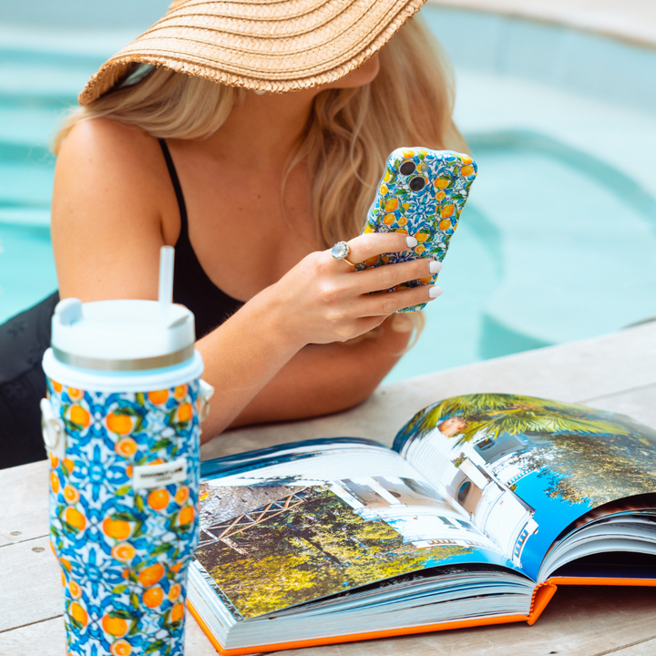 A woman in a sun hat and black swimsuit holds a smartphone with a colorful case while sitting by the pool. In front of her is a large, tropical-themed Stanley Quencher placed inside the Coconut Lane Tumbler Carry Case - Amalfi Lemon and an open vacation-themed magazine.