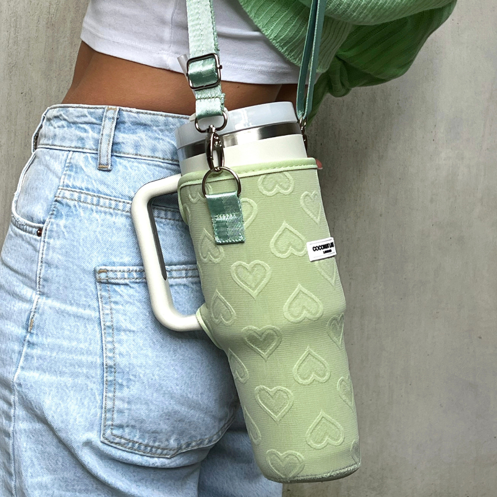 A person wearing light blue jeans and a green shirt carries a travel mug with a heart pattern in a matching Coconut Lane Tumbler Carry Case - Sage Heart, slung across their shoulder. The case has both a handle and strap for easy carrying.