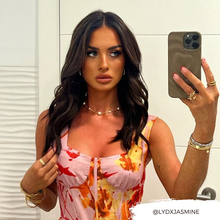A woman with long, dark hair takes a mirror selfie with her smartphone covered by the Coconut Lane Melting Heart Phone Case in Chocolate Brown. She is wearing a colorful dress adorned with red and yellow patterns, along with several accessories such as necklaces, bracelets, and rings. Her expression is neutral. Displayed on the image is the Instagram handle @lydxjasmine.