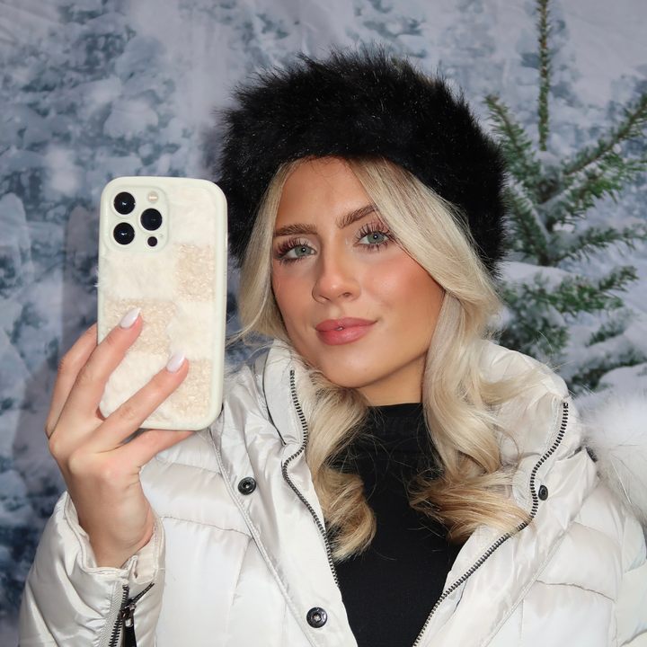A person with long blonde hair takes a selfie with their smartphone, which is encased in the Cosy Teddy Phone Case - Vanilla Biscuit from Coconut Lane. They are wearing a white quilted jacket and a black fur hat, standing against a snowy winter background with trees.