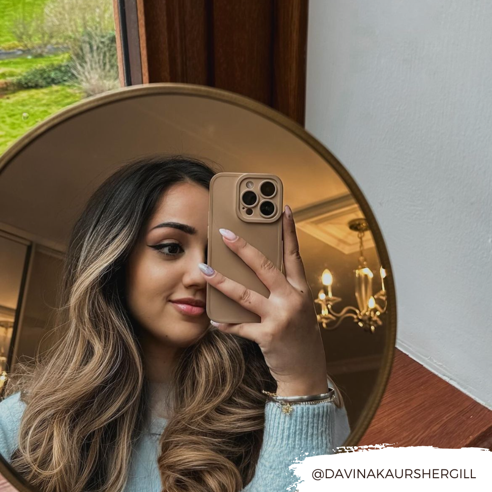 A woman with long, wavy hair takes a mirror selfie inside a room with wooden furnishings. She is holding a beige smartphone adorned with the NAKD Phone Case - Peach by Coconut Lane and wearing a light blue sweater. A chandelier is visible in the background. The Instagram handle "@DAVINAKAURSHERGILL" is written at the bottom right.