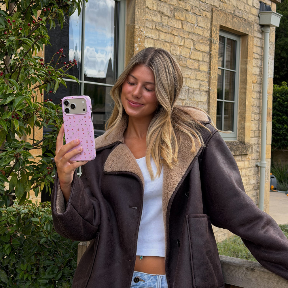 Woman wearing brown aviator jacket holds up an iphone in a baby pink phone case, with small dainty pastries, pumpkins, wooly hats, bows and leaves printed across the case.