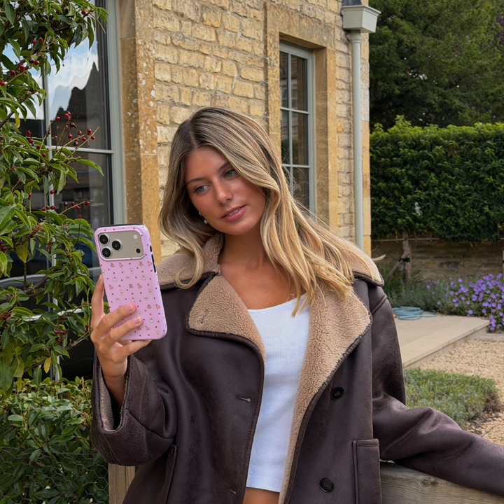 Woman wearing brown aviator jacket holds up an iphone taking a selfie, with a baby pink phone case, with small dainty pastries, pumpkins, wooly hats, bows and leaves printed across the case.