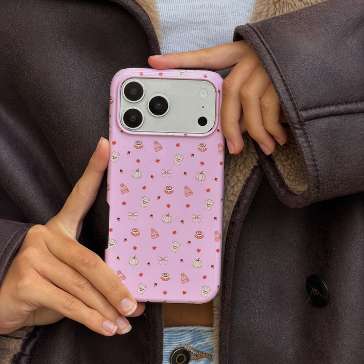 Woman wearing brown aviator jacket holds up an iphone in a baby pink phone case, with small dainty pastries, pumpkins, wooly hats, bows and leaves printed across the case.