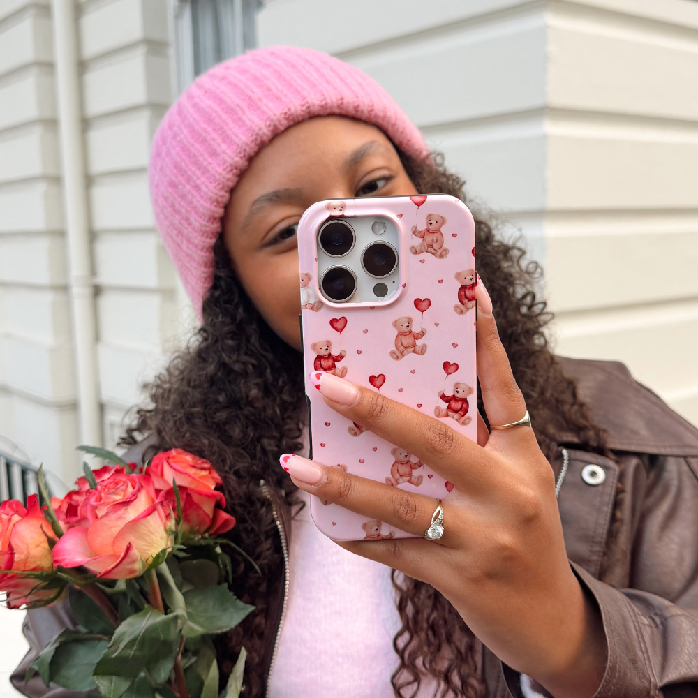 Person taking a selfie on a phone with a pink case featuring teddy bears and hearts, wearing a pink beanie and holding flowers.