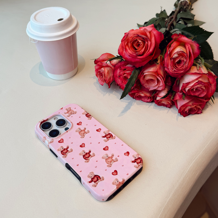 Phone case with teddy bear and heart pattern next to red roses and a coffee cup on a light surface.