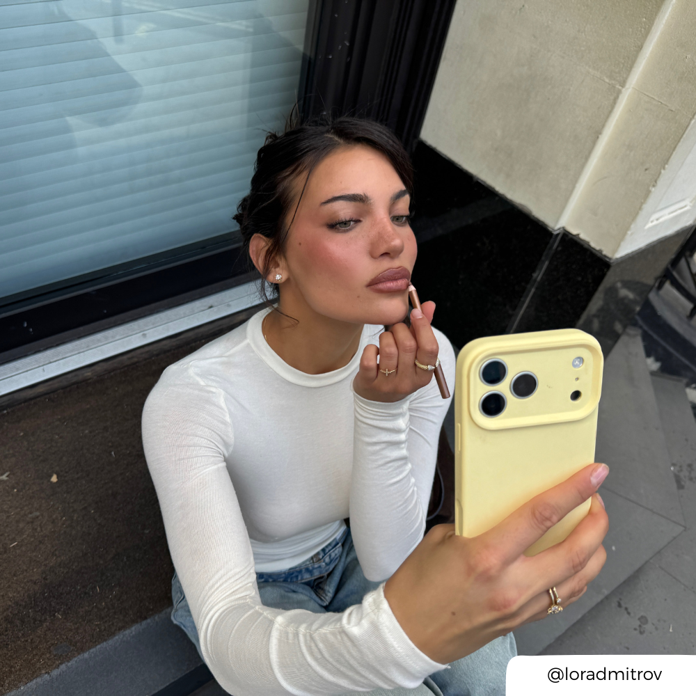 a woman applying makeup in a street setting, holding an iphone in a yellow pastel phone case.