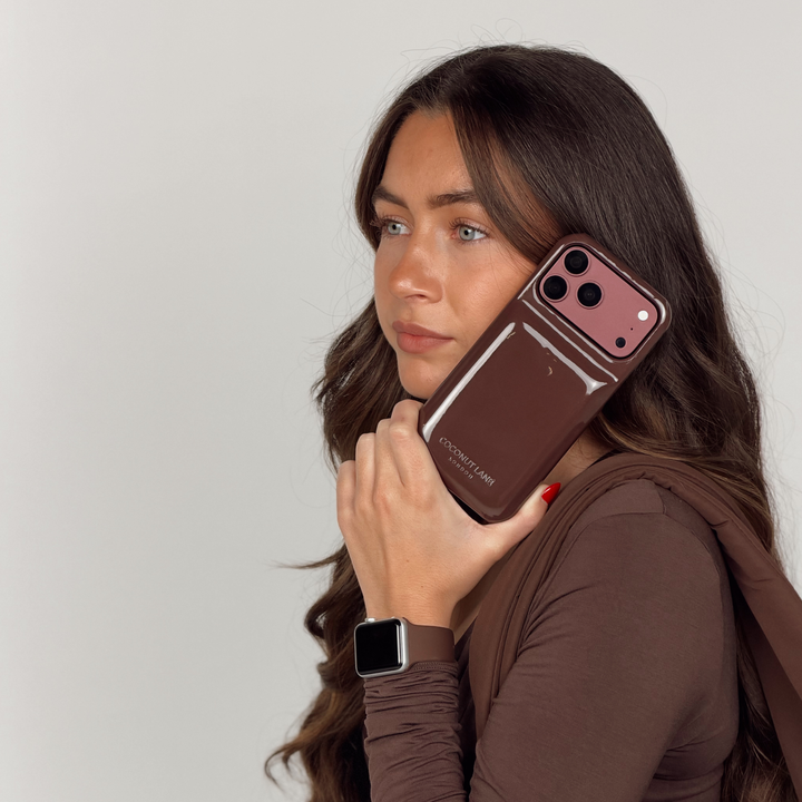 Woman wearing a brown pilates outfit, holding an iphone up to her ear. The phone is in a glossy chocolate brown Coconut Lane puffer phone case with embossed logo. On her wrist is a Coconut Lane NAKD Chocolate brown apple watch strap.