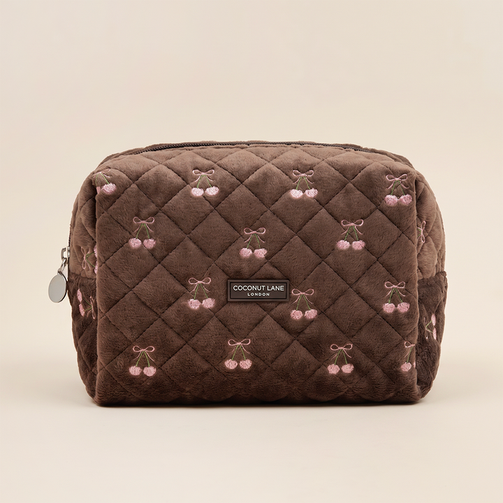 Brown quilted velvet makeup bag with pink embroidered cherries tied with bows and a “Coconut Lane London” label on the front.