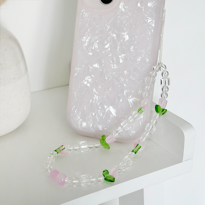 A smartphone with a textured, pearlescent white case is placed on a white surface. Attached to the phone is the "Beaded Phone Strap - Cute Tulips" by Coconut Lane, made of clear, pink, and green beads, which rests neatly on the same surface. A small portion of a plant pot is visible in the corner.