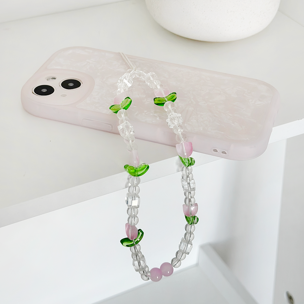 A white smartphone with a marble-patterned case lies on a white surface. Draped over it is the Beaded Phone Strap - Cute Tulips from Coconut Lane, featuring transparent, green, and purple beads arranged in a stylish pattern. This chic phone accessory sits on the edge of the surface, adding a decorative touch.