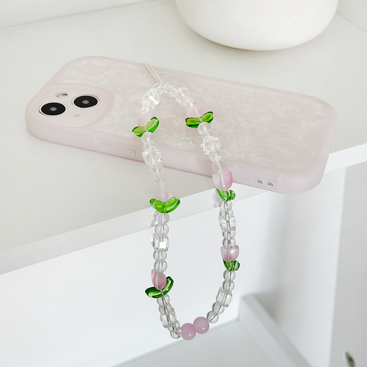 A white smartphone with a marble-patterned case lies on a white surface. Draped over it is the Beaded Phone Strap - Cute Tulips from Coconut Lane, featuring transparent, green, and purple beads arranged in a stylish pattern. This chic phone accessory sits on the edge of the surface, adding a decorative touch.