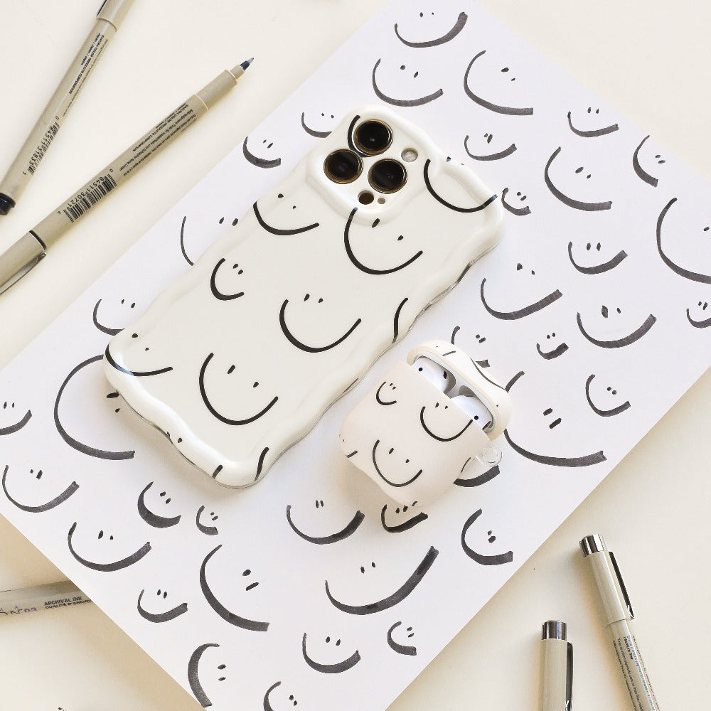 A Wavy Phone Case - Smiley Doodle by Coconut Lane and an AirPods case, both featuring a minimalist black smiley doodle design on a white background, are placed on a matching sheet of paper. Surrounding the sheet are several black ink pens. The Coconut Lane Wavy Phone Case adds an extra protective touch to this charming setup.