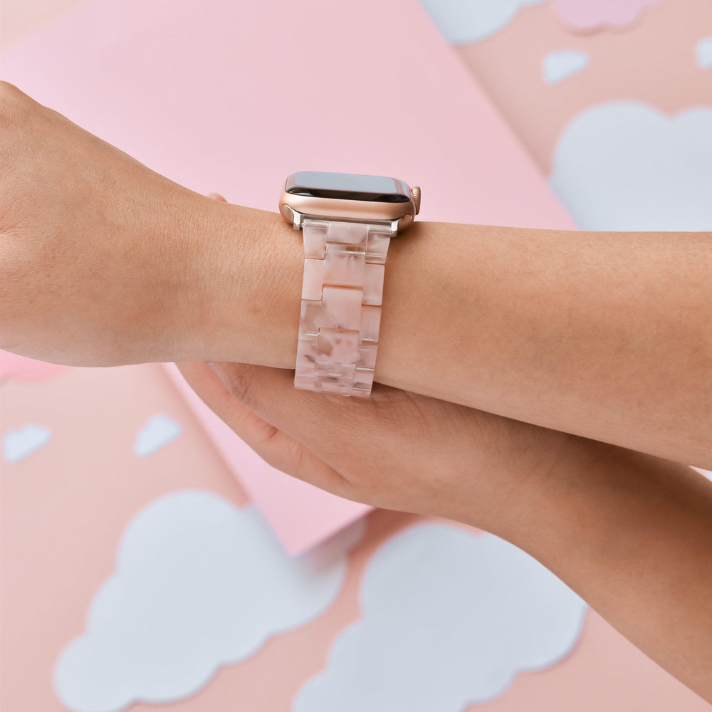 Close-up of a person wearing an Apple Watch with the Luxe Pink Clouds Apple Watch Strap from Coconut Lane on their wrist. The background features a soft pastel-colored design with pink and white paper clouds, highlighting the strap's durable resin craftsmanship.