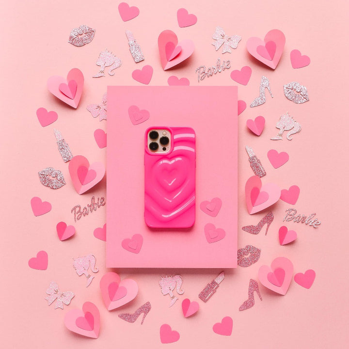A bright pink iPhone, adorned with Coconut Lane's Melting Heart Phone Case, lies on a pink surface surrounded by small pink paper hearts and an array of glittery decorations such as shoes, lipsticks, and the word "Barbie." The scene exudes a playful and feminine aesthetic.