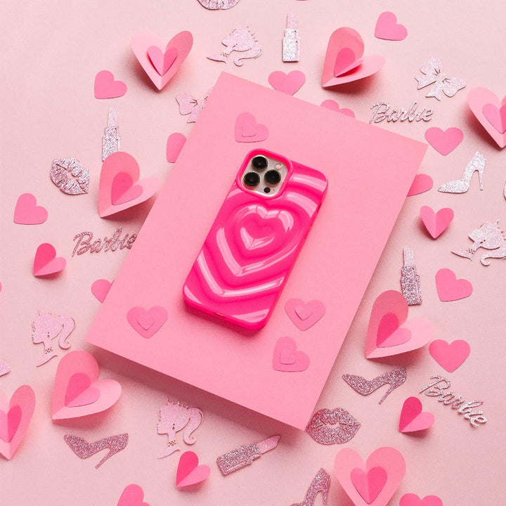 A Coconut Lane "Melting Heart Phone Case - Bright Pink" smartphone is placed on a light pink rectangular surface. Surrounding the phone are various pink and glittery cutouts of hearts, unicorns, high heels, and the word "Barbie." The background is a soft pink.