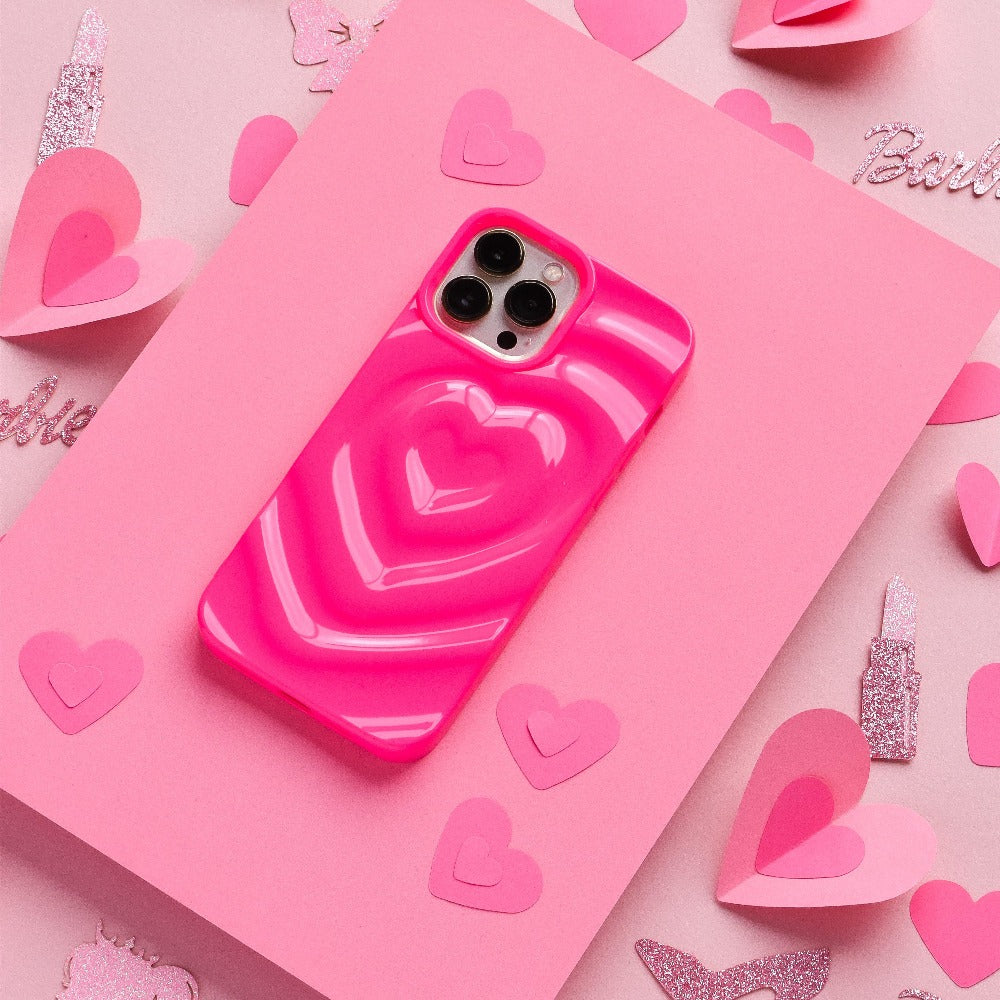 A Coconut Lane smartphone, adorned with a Melting Heart Phone Case - Bright Pink featuring 3D melting heart effects, is placed on a pink sheet of paper. The background is embellished with pink paper hearts and glittery cutouts, creating a vibrant and cheerful setting.