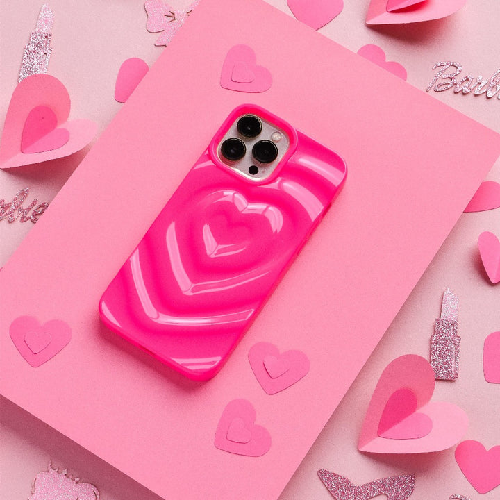A Coconut Lane smartphone, adorned with a Melting Heart Phone Case - Bright Pink featuring 3D melting heart effects, is placed on a pink sheet of paper. The background is embellished with pink paper hearts and glittery cutouts, creating a vibrant and cheerful setting.