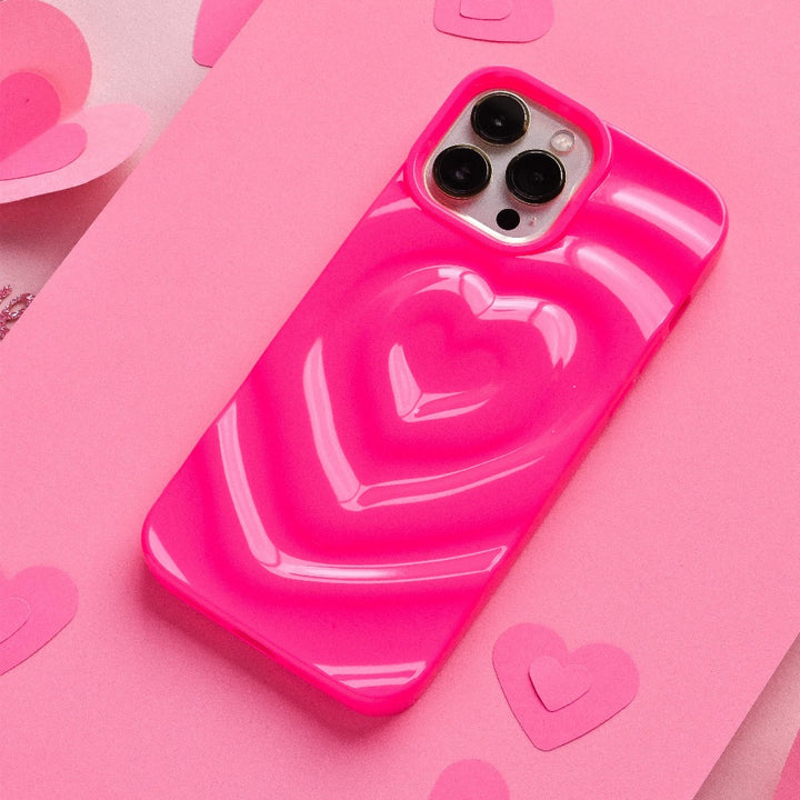 The Melting Heart Phone Case - Bright Pink by Coconut Lane, featuring a raised heart design with a 3D melting heart effect, is lying on a pink surface. The extra protective phone case is surrounded by small paper hearts, enhancing the overall pink-themed, romantic aesthetic.